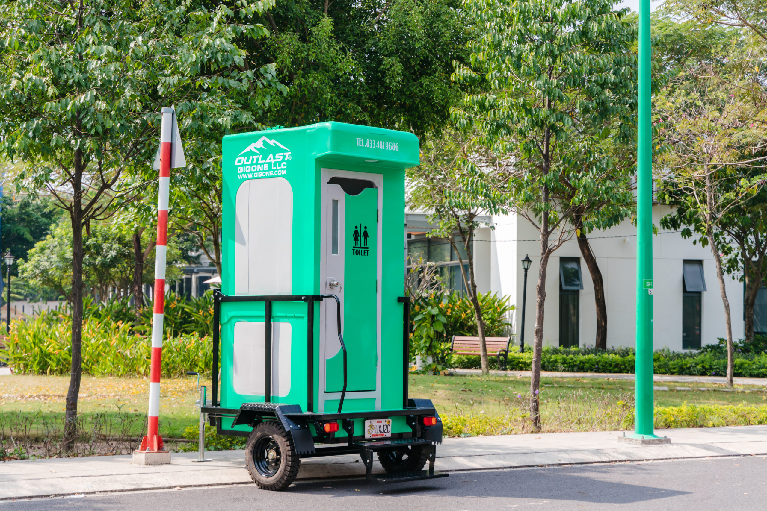 Gigone LLC portable toilet unit on a wheeled trailer, parked along a tree-lined street in a park setting